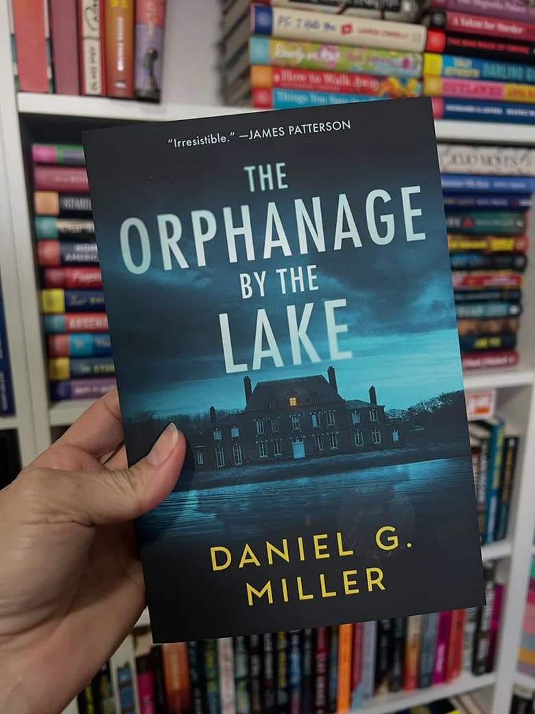 A reader holding The Orphanage by the Lake by Daniel G. Miller in front of bookshelves